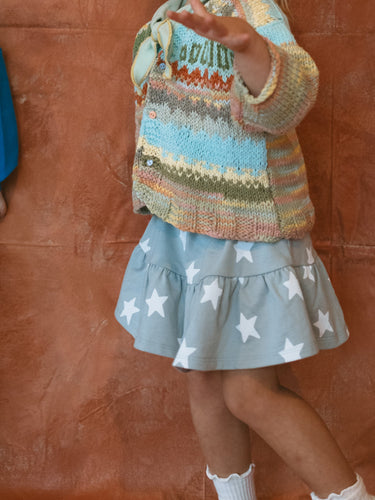 Two children standing against a textured brown wall, one wearing a colorful sweater and star-patterned skirt.