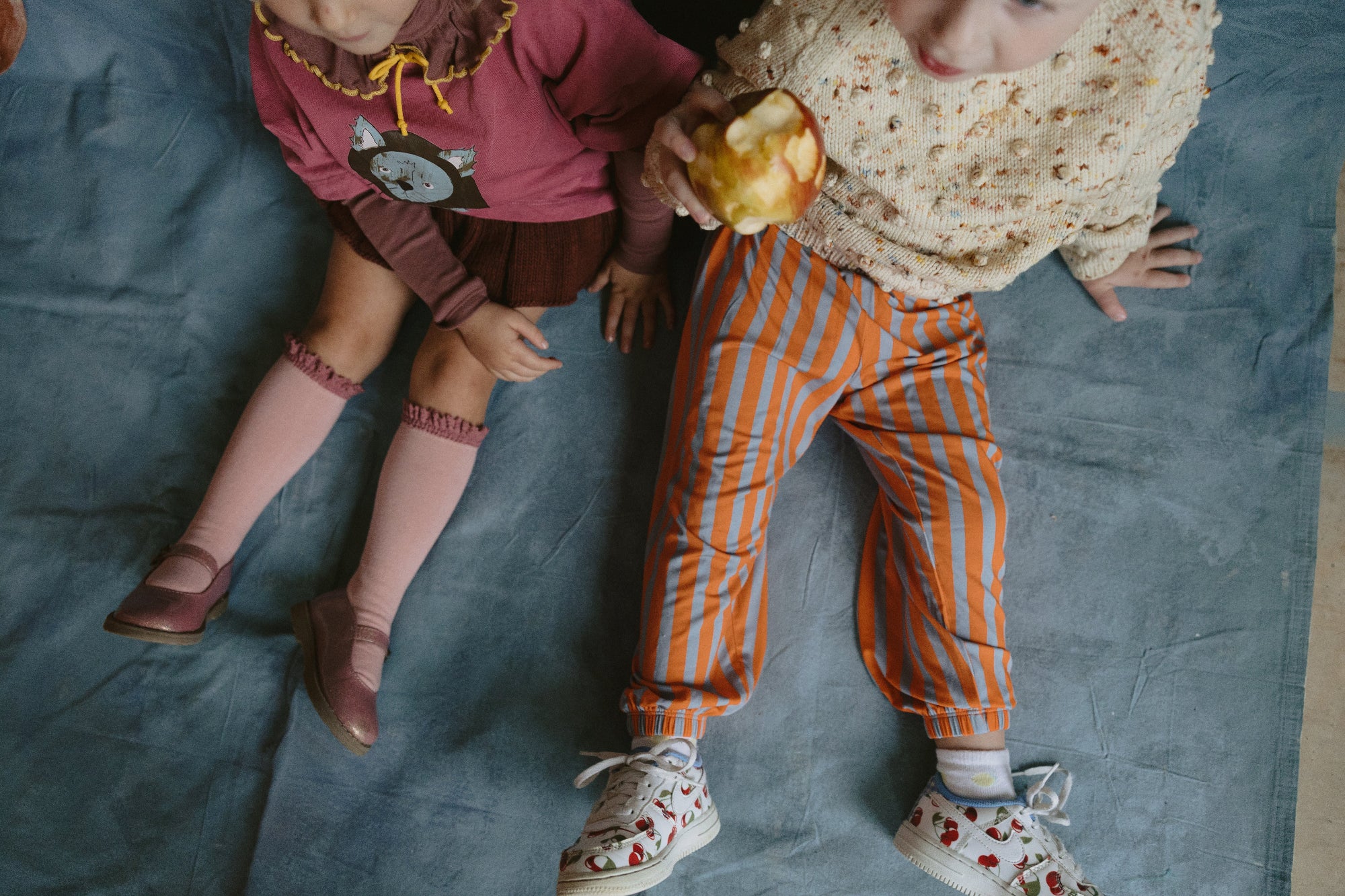 Two children in colorful outfits on a blue blanket, one eating an apple.