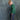 A young child standing in profile against a mottled blue-gray studio backdrop, wearing a green striped long-sleeve shirt and dark navy pants with a white paisley print down the side.