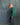 A young child standing in profile against a mottled blue-gray studio backdrop, wearing a green striped long-sleeve shirt and dark navy pants with a white paisley print down the side.