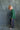 A young child standing in profile against a mottled blue-gray studio backdrop, wearing a green striped long-sleeve shirt and dark navy pants with a white paisley print down the side.