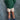 A young child standing against a mottled blue-gray studio backdrop, wearing a green and dark green striped long-sleeve shirt, cream shorts with black polka dots, blue socks, and purple sneakers.