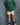 A young child standing against a mottled blue-gray studio backdrop, wearing a green and dark green striped long-sleeve shirt, cream shorts with black polka dots, blue socks, and purple sneakers.