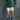 A child wearing a green and dark green striped long-sleeve shirt, cream shorts with black polka dots, blue and white striped socks, and purple sneakers, standing against a mottled gray backdrop.