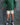 A child wearing a green and dark green striped long-sleeve shirt, cream shorts with black polka dots, blue and white striped socks, and purple sneakers, standing against a mottled gray backdrop.