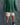 A child wearing a green and dark green striped long-sleeve shirt, cream shorts with black polka dots, blue socks, and purple sneakers, standing against a mottled gray studio backdrop.